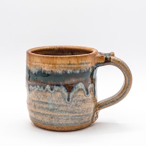 May include: Handmade ceramic mug with a brown, blue, and tan glaze. The mug features a rounded handle and a textured surface. The glaze has a dripped effect, adding to its unique, rustic appearance. A perfect addition to any kitchen.