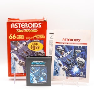 May include: A vintage Atari Asteroids video game set. The set includes a red box with the game title and a spaceship graphic, a game cartridge, and an instruction manual. The box has a "SALE! $9.99" sticker.