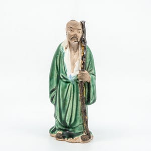 May include: A ceramic figurine of a person with a shaved head and a mustache, wearing a long green robe with a white collar. The figure holds a tall, dark brown, textured staff in the left hand. The base and skin tones are a light tan color.