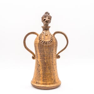 May include: A handcrafted, brown ceramic pitcher with two curved handles. The pitcher features a decorative face on the stopper and a sunburst design. The ceramic piece is approximately 12 inches tall.