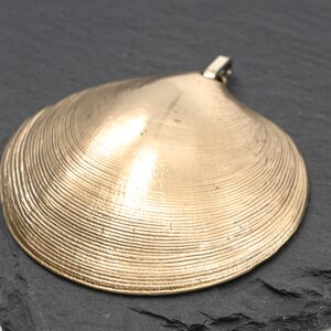 May include: A gold-toned pendant shaped like a clam shell, featuring a textured surface with concentric ridges and a small metal loop at the top for attachment. The object has a smooth, metallic finish and a curved, semi-circular silhouette.