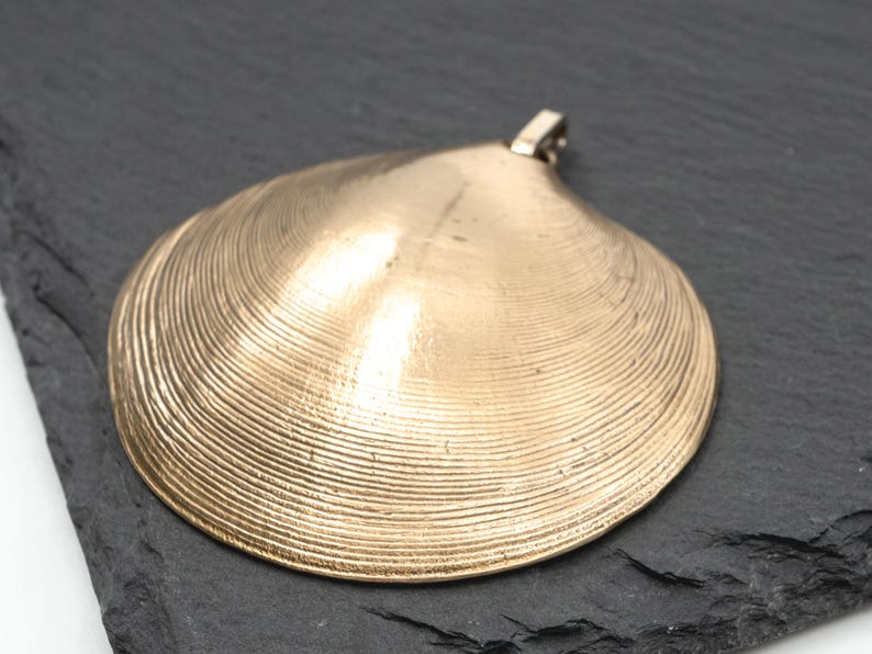 May include: A gold-toned pendant shaped like a clam shell, featuring a textured surface with concentric ridges and a small metal loop at the top for attachment. The object has a smooth, metallic finish and a curved, semi-circular silhouette.