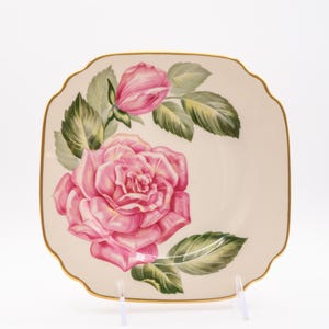 May include: Square, off-white plate with a pink rose and bud design. The rose is in full bloom with green leaves. The plate has a gold trim and a scalloped edge. The plate is on a clear stand.