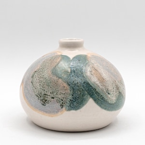 May include: A small, round ceramic vase with a narrow neck. The vase is primarily off-white with abstract, flowing patterns in shades of blue, green, and tan. The design gives the vase a unique, artistic appearance.
