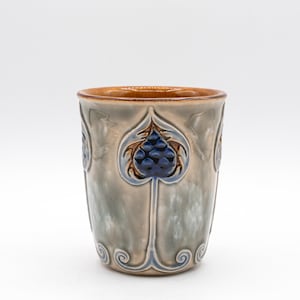 May include: A ceramic mug with a light gray and blue glaze. The mug features a repeating pattern of stylized blue floral designs. The rim is brown, and the interior is a lighter brown. The mug is cylindrical and appears to be handmade.
