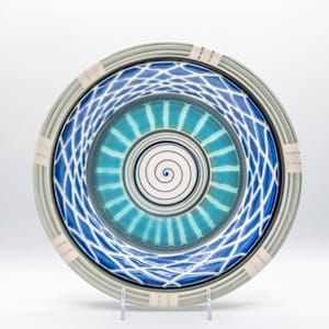 May include: A decorative ceramic plate with a blue and white geometric design. The plate features concentric circles of color, including a central spiral, surrounded by blue and white patterns. The rim has a gray and white striped pattern.