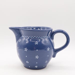 May include: A dark blue ceramic pitcher with a rounded body, a spout, and a handle. The pitcher is decorated with small white floral patterns and a white zigzag pattern around the rim. The pitcher is approximately 7 inches tall.