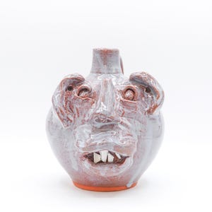 May include: A ceramic jug with a face design. The jug is primarily gray with red and white accents, featuring a face with protruding ears, eyes, and teeth. The base is orange, and the jug has a handle.