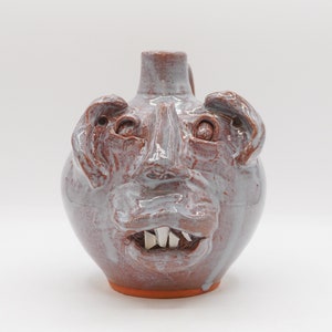 May include: A ceramic jug with a face sculpted on the front. The jug is glazed in a light blue color with brown accents. The face has large eyes, a wide mouth, and a row of teeth.