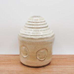 May include: A beige ceramic vase with a textured surface and a rounded shape. The top portion features horizontal ridges, while the lower part has three circular indentations. The vase sits on a wooden surface.