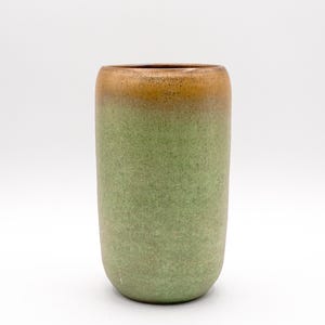 May include: A cylindrical ceramic vase with a gradient color scheme. The top rim is a warm brown, transitioning to a light green body. The vase has a textured surface and a rounded base, suitable for home decor.