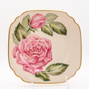 May include: Square, off-white plate with a pink rose and bud design. The rose is in full bloom with detailed petals, surrounded by green leaves. The plate has a gold trim and a scalloped edge, creating an elegant look.