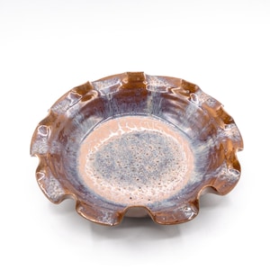 May include: A round ceramic bowl with a ruffled, scalloped edge. The interior features a textured, speckled finish in shades of light pink and grey, transitioning into a glossy, deep brown glaze that extends to the outer rim and wavy border.