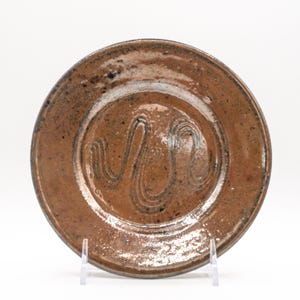 May include: A handmade, rustic ceramic plate with a brown glaze and a white interior. The plate features a textured, wavy design in the center. The plate is displayed on a clear stand against a white background.