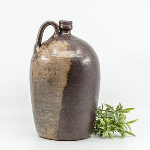 May include: A stoneware jug with a rounded body, a narrow neck, and a single handle. The exterior features a two-tone glaze with a dark brown finish on one side and a lighter, mottled tan and grey texture on the other side of the vessel.