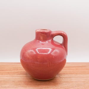May include: A small, red ceramic vase with a rounded body and a single handle. The vase has a glossy finish.