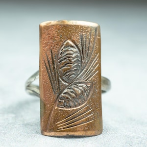 May include: A rectangular copper-colored ring with a silver band. The copper face features an embossed design of two pine cones and needles. The ring is a unique piece of jewelry, suitable for everyday wear or special occasions.