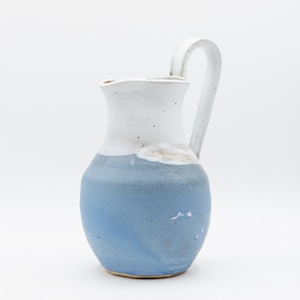 May include: A handmade ceramic pitcher with a curved handle. The bottom half is a light blue, transitioning to white at the top. The pitcher has a rustic, textured appearance, suitable for serving beverages.