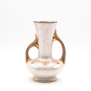 May include: A ceramic vase with an iridescent pearl finish and two gold-toned handles. The body features a rounded base and a flared rim, decorated with a repeating gold floral pattern around the center. The rim and base are trimmed in gold.