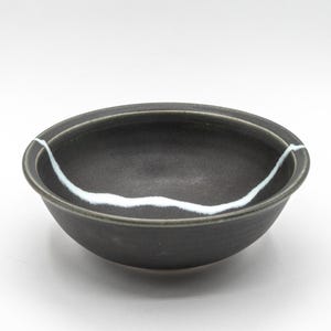 May include: A dark gray ceramic bowl with a white, wavy line accent. The bowl has a simple, rounded shape and a smooth, matte finish. The white line appears to be a decorative element or a repair.
