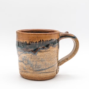 May include: Handmade ceramic mug with a unique, textured design. The mug features a brown rim, a dark blue and brown band, and a tan body. The handle is curved and matches the mug's color scheme. A rustic, artisanal piece.