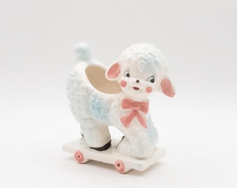 May include: A vintage ceramic lamb planter in white, light blue, and pink. The lamb has a textured coat, pink ears, and a bow. It is mounted on a white skateboard with pink wheels. The planter has an opening on its back.