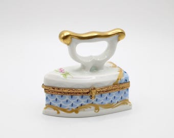 May include: A decorative porcelain trinket box shaped like an antique iron. The white iron features a gold handle and trim, with a blue and gold patterned base. Delicate floral accents add a touch of color to this charming collectible.