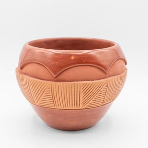 May include: A handcrafted terracotta-colored ceramic bowl. The bowl features a decorative band of carved geometric patterns around the middle and a scalloped design near the rim. The bowl has a smooth, glossy finish.