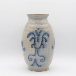 May include: A beige ceramic vase with blue floral designs. The vase has a wide body and a narrow neck. The blue design is symmetrical and covers the front of the vase. The vase is on a white background.