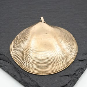 May include: A gold-toned metal pendant shaped like a clam shell, featuring a textured surface with concentric ridges and a small metal loop at the top for attachment. The pendant has a smooth, metallic finish and a distinct triangular shape.