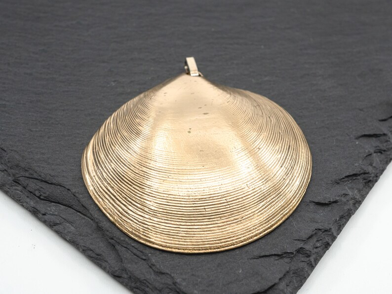 May include: A gold-toned metal pendant shaped like a clam shell, featuring a textured surface with concentric ridges and a small metal loop at the top for attachment. The pendant has a smooth, metallic finish and a distinct triangular shape.
