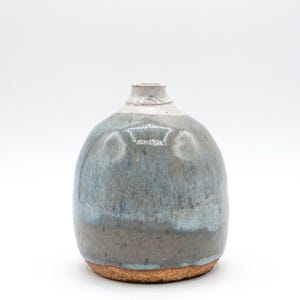 May include: A small, rounded ceramic vase with a narrow neck. The vase features a gradient of colors, transitioning from a light gray at the top to a blue-gray in the middle and a darker gray at the base. The base has a textured, brown appearance.
