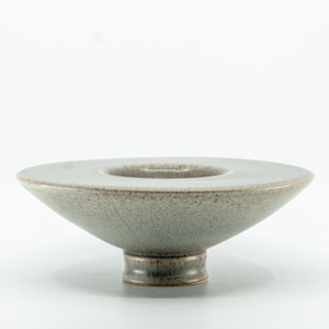 May include: A wide, shallow ceramic bowl with a tapered, pedestal-style base. The exterior features a speckled, mottled glaze in shades of muted grey, beige, and brown, with a smooth, light-colored interior surface and a defined circular rim.