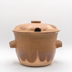 May include: A brown ceramic casserole dish with a lid. The dish has two handles and a small opening in the lid. The exterior features a darker brown glaze that drips down the sides. The dish is designed for cooking and serving.