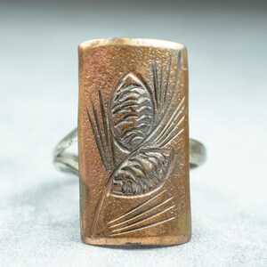 May include: A rectangular copper-colored ring with a silver band. The copper face features an embossed design of two pine cones and needles. The ring is set against a light gray background.