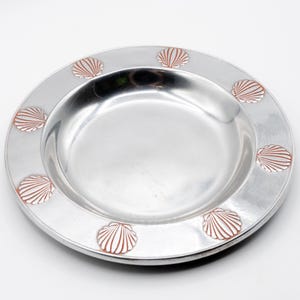May include: A silver-colored plate with a wide rim, decorated with embossed seashell designs in a reddish-brown color. The plate has a shallow, concave center and is likely made of metal. The seashell pattern is evenly spaced around the rim.