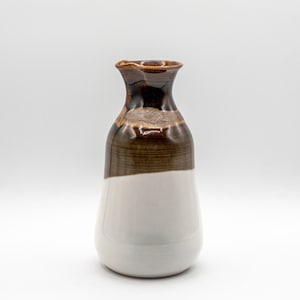 May include: A ceramic vase with a unique design. The top is dark brown, transitioning to a lighter brown band, and then to a white base. The vase has a slightly flared, textured rim.