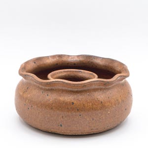 May include: A handmade, brown ceramic candle holder with a wavy rim and a central circular well. The pottery has a speckled texture and a rustic appearance, perfect for home decor.