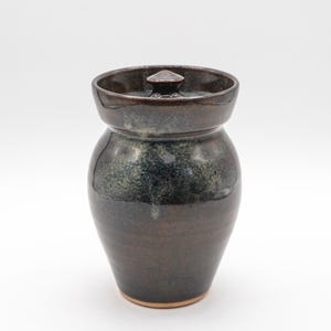 May include: A handcrafted, dark brown ceramic jar with a lid. The jar has a rounded body and a slightly flared rim. The lid features a small, pointed handle. The glaze has a speckled, dark green and brown appearance.