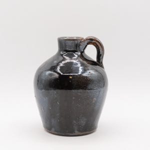 May include: A small, dark brown ceramic jug with a handle. The jug has a rounded body and a narrow neck. The surface has a glossy finish with some imperfections. The handle is arched and attached to the neck and body.