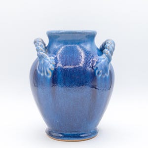 May include: A blue ceramic vase with a glossy finish. The vase has two rope-like handles and a wide, rounded body. The color is a deep, rich blue, with subtle variations in tone. The vase is a decorative item.