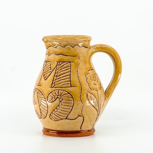 May include: A tan ceramic pitcher with a glossy finish, featuring a curved handle and a scalloped rim. The exterior is decorated with the etched letters M and O, along with a floral design on the side, all rendered in a dark brown outline.