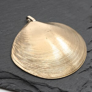 May include: A gold-toned metal pendant shaped like a clam shell, featuring a detailed ribbed texture across its surface and a small metal loop at the top for attachment. The pendant has a smooth, reflective finish with visible concentric lines.
