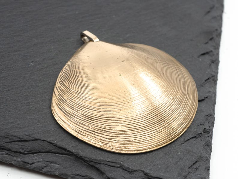 May include: A gold-toned metal pendant shaped like a clam shell, featuring a detailed ribbed texture across its surface and a small metal loop at the top for attachment. The pendant has a smooth, reflective finish with visible concentric lines.