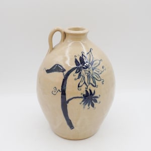 May include: A beige ceramic jug with a handle and a blue floral design. The hand-painted flower has multiple petals and is centered on the jug's body. The jug has a rounded shape and a small opening at the top.