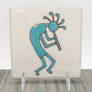 May include: A turquoise blue ceramic tile with a stylized Native American flute player design. The tile is on a white background and has a brown outline.