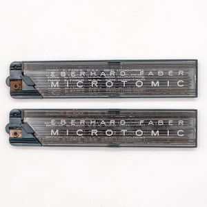 May include: Two rectangular, dark gray pencil lead holders with the text "EBERHARD FABER MICRO-TOMIC" printed on them. The holders are made of a translucent material, and the leads are visible inside. The holders are on a white background.