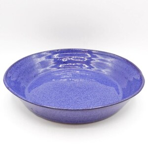 May include: A round ceramic bowl with a vibrant purple glaze featuring a speckled texture. The bowl has a wide, shallow basin with gently sloping sides and a smooth, glossy finish that reflects light across its curved interior surface.