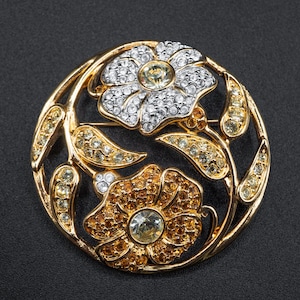 May include: A gold-toned brooch featuring two flower designs with crystal accents. The brooch has a circular shape with a floral and leaf design. The flowers are white and amber with a central crystal. The leaves are amber-colored.