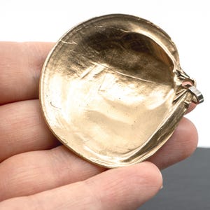 May include: A gold-toned metal pendant shaped like a rounded seashell. The surface has a smooth, reflective finish with subtle indentations and a metallic sheen. A small silver-colored metal loop is attached to the side for hanging or attachment.